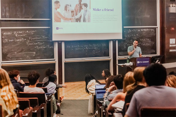 纽约大学柯朗学院研究生新生欢迎会现场,讲者正在向新生介绍校园生活与社交建议,背景黑板上展示数学公式,体现学院浓厚的学术氛围与人文关怀。